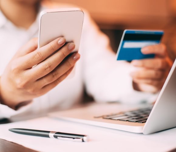 Close-up of female hand holding smartphone. Young businesswoman sitting at laptop in office holding credit card and entering data on her mobile phone